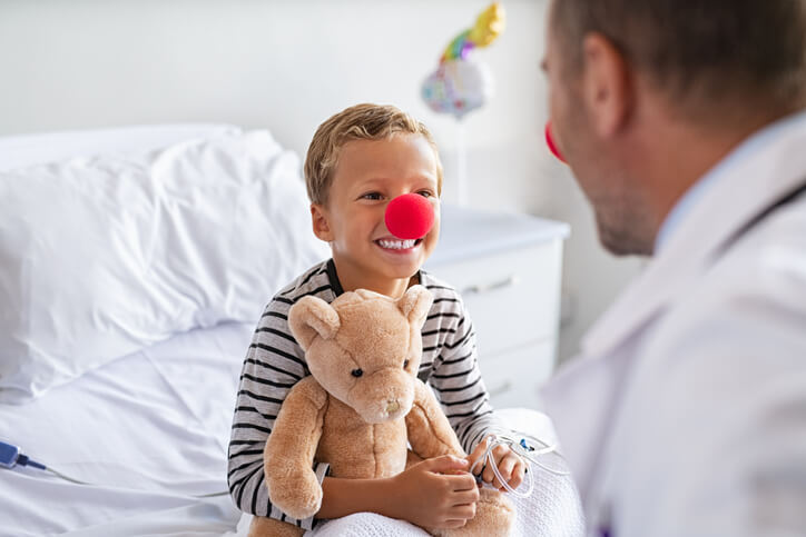Lachende jongen met rode clownneus op ziekenhuisbed houdt een knuffel vast en kijkt naar een CliniClown – symbool voor de impact van donaties aan CliniClowns.